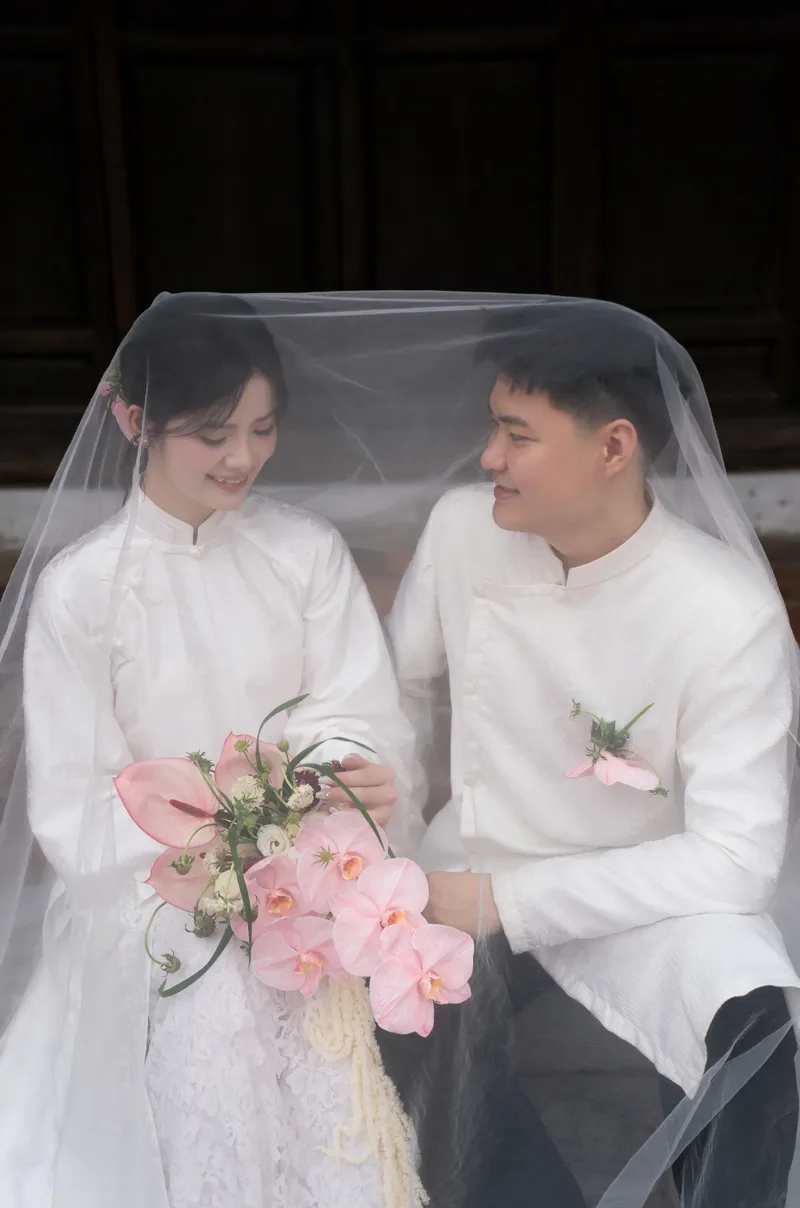 Cô dâu chú rể mặc áo dài trắng dưới tấm voan, cô dâu cầm hoa hồng