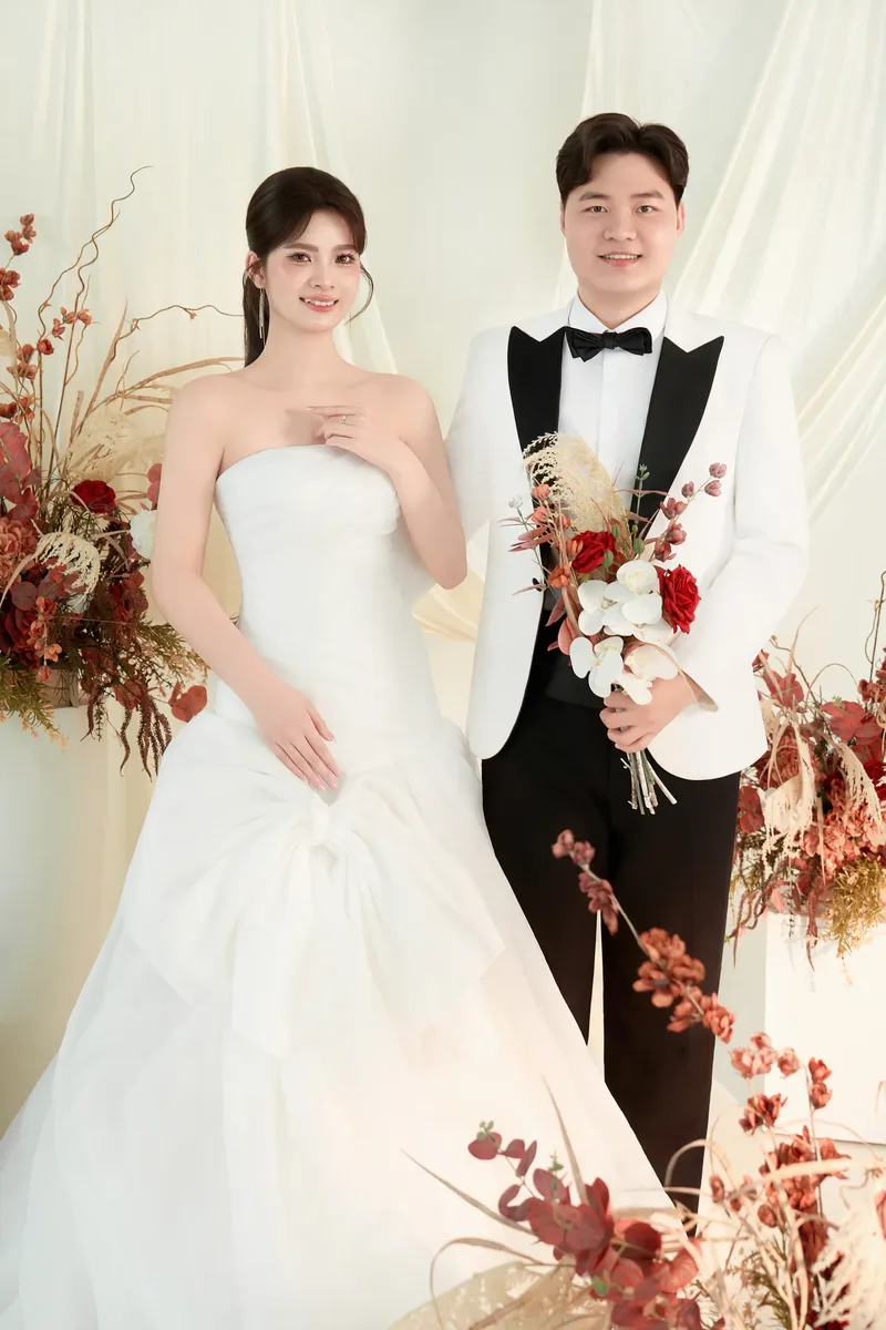 Cô dâu chú rể bên những cành hoa đỏ trên nền rèm kem