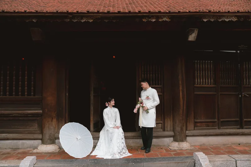 Cô dâu ngồi cạnh chiếc ô giấy trắng, chú rể cầm hoa đứng trước ngôi nhà gỗ cổ mái ngói đỏ